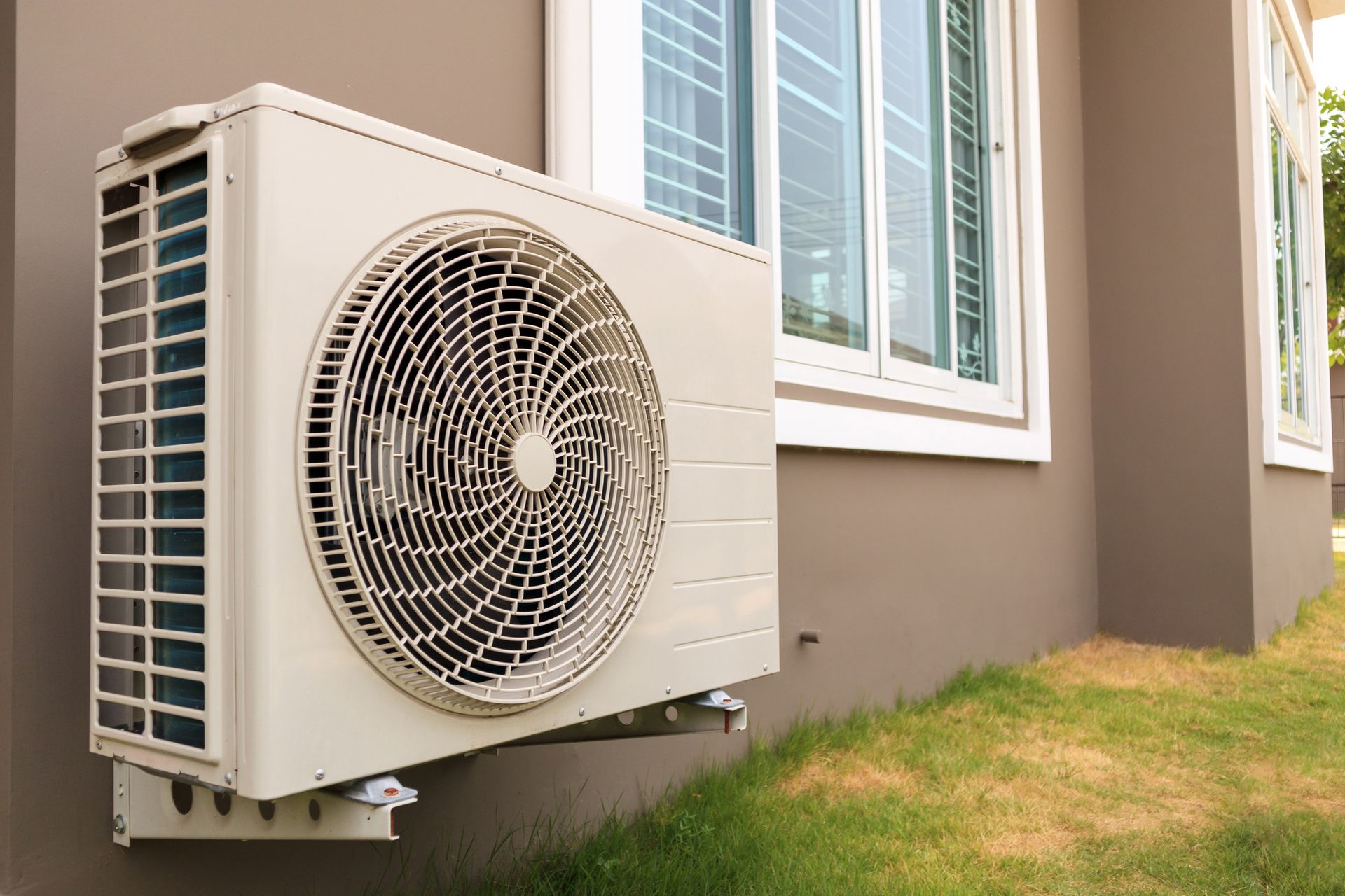 An air conditioner is mounted on the side of a house next to a window.