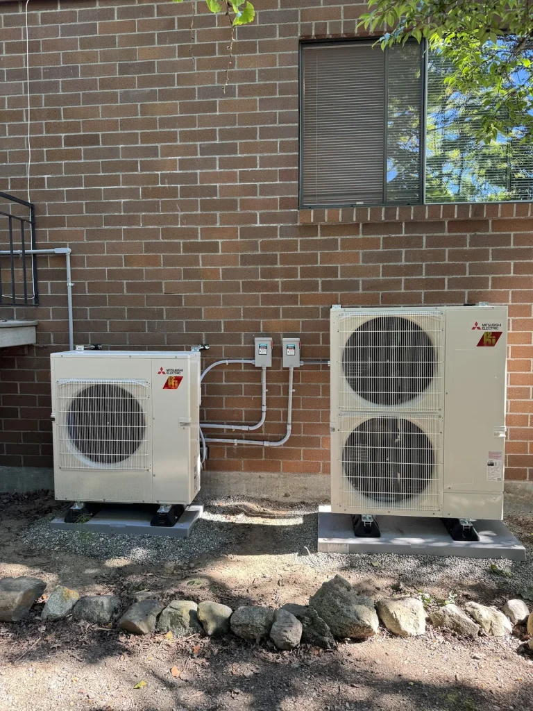 Two Mitsubishi outdoor units installed on pads beside a brick building